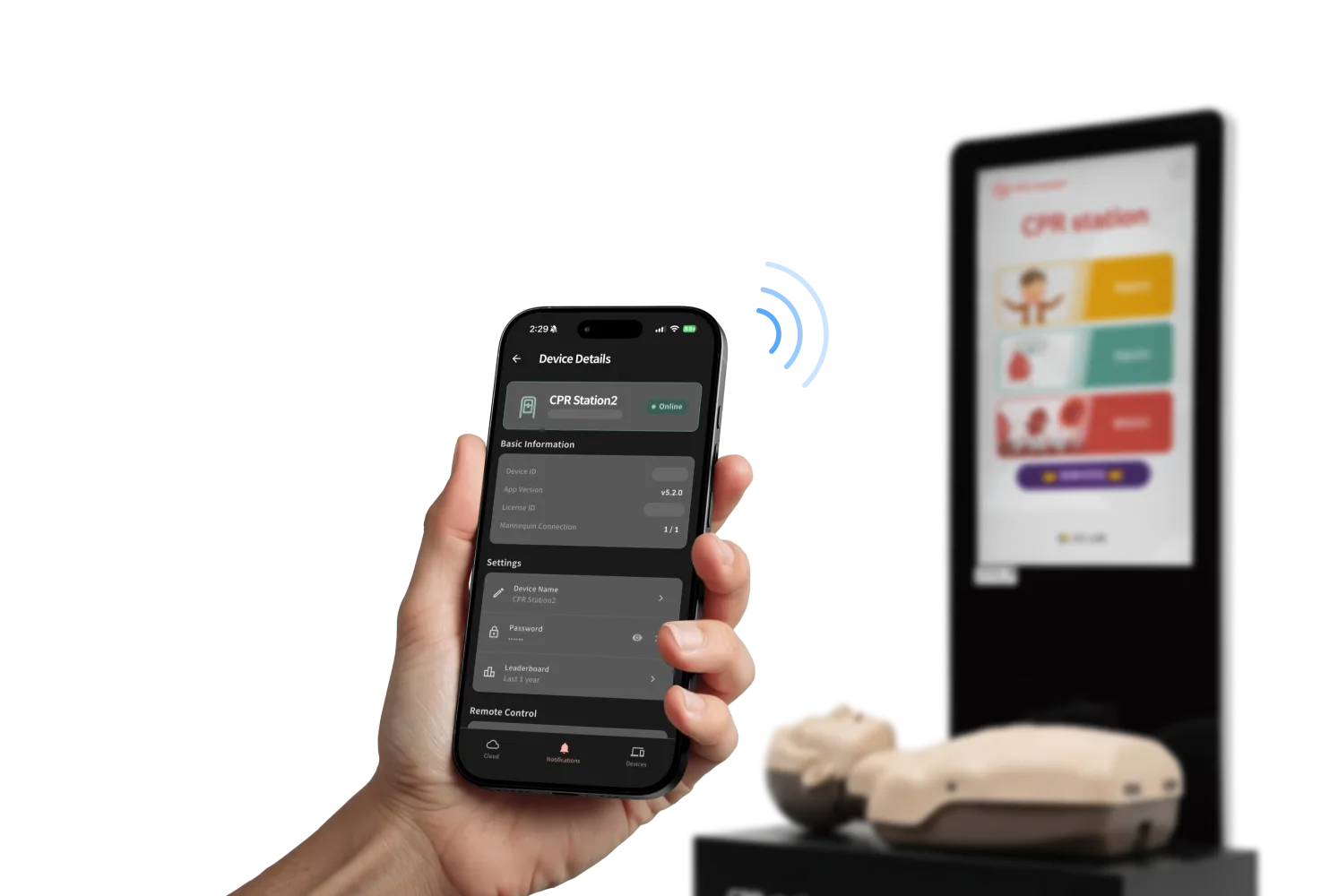 A phone showing the CPR cloud app's device details screen — kiosk name, Online status, app version, and manikin connection count — wirelessly linked to a CPR station kiosk standing behind it.