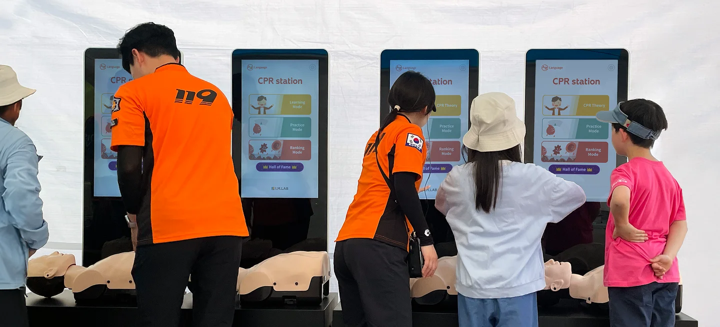 Visitors and paramedics practicing on four CPR station kiosks at an outdoor event